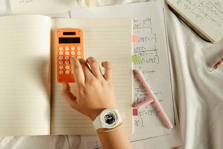 Eine Person tippt auf einem orangen Taschenrechner, darunter liegen Hefte mit mathematischen Formeln. Die Peterssche Formel ist eine Möglichkeit, um die Höhe der Instandhaltungsrücklage bei Immobilien zu ermitteln.