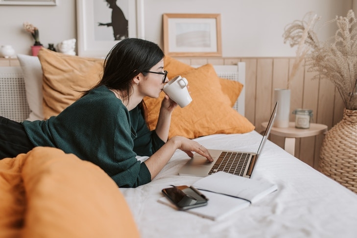 Eine Frau liegt auf einem Bett und schaut auf ihren Laptop, während sie Kaffee trinkt. In Zukunft könnten Ihre Teilnahme an der Eigentümerversammlung genau so aussehen. Dazu brauchen Sie allerdings auch die technischen Voraussetzungen, um an Videocalls teilzunehmen.