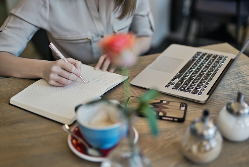 Eine Frau sitzt vor ihrem Laptop und macht sich Notizen, vor ihr steht eine Tasse Kaffee.